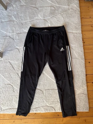 Svarta Adidas träningsbyxor - Säljer ett par svarta träningsbyxor från Adidas med klassiska vita ränder längs sidorna och logga på vänster lår. Byxorna har smal passform, resår i midjan och är tillverkade i ett lätt och stretchigt material som passar perfekt till träning.