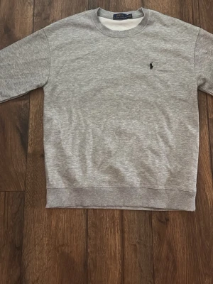 Grå sweatshirt från Polo Ralph Lauren - Klassisk grå sweatshirt från Polo Ralph Lauren med rund halsringning och den ikoniska svarta logotypen broderad på bröstet. Tröjan har ribbade muddar vid ärmslut och nederkant. Perfekt för en avslappnad och stilren look.
