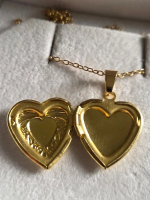 Guldigt hjärtformat medaljonghalsband - Snyggt halsband med hjärtformat medaljonghänge i guldfärg. Medaljongen har fina blommönster och en liten rosett på framsidan. Kedjan är tunn och elegant i rostfritt stål och medaljongen går att öppna för att lägga in ett litet foto eller minne.