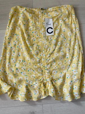 Gul blommig kjol från Cubus - Söt gul kortkjol från Cubus med vitt och grönt blommönster. Kjolen har volangkant nertill och en snygg rynkning framtill som ger extra detalj. Perfekt för vår och sommar med sitt lätta och luftiga material.