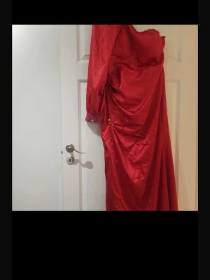Röd långklänning med pärldetaljer - Snygg röd långklänning med en axel och lång ärm. Klänningen har pärldetaljer vid ärmslutet och draperad effekt längs sidan. Perfekt för dig som vill sticka ut på bal eller fest. Materialet har en glansig finish.