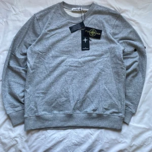 Grå stickad sweatshirt Stone Island - Säljer en grå stickad sweatshirt från Stone Island med klassisk rund hals och ribbade muddar. Tröjan har en enkel design med logotyp på ärmen och är perfekt för dig som gillar stilrena plagg. Passar till många olika outfits.