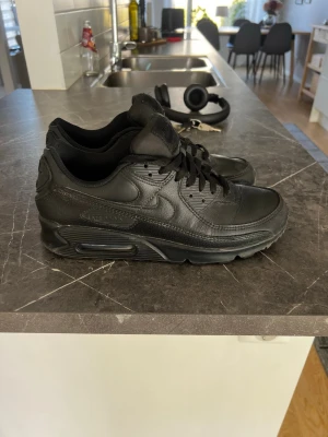 Nike Air Max 90 helsvarta sneakers - Säljer ett par klassiska Nike Air Max 90 sneakers i helsvart läder och syntet. Skorna har den ikoniska Air Max-sulan, svarta skosnören och diskreta detaljer som ger en clean look. Perfekta för dig som gillar stilrena och tidlösa sneakers.