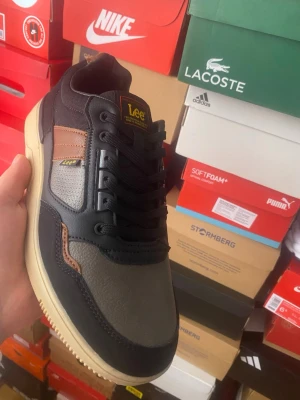 Lee sneakers i svart och grått - Snygga sneakers från Lee i svart och grått med bruna detaljer. Ovandelen är i en mix av läder och syntetmaterial, med svarta skosnören och beige sula. Klassisk låg modell med Lee-logga på sidan och plös. Perfekta för en stilren streetlook.