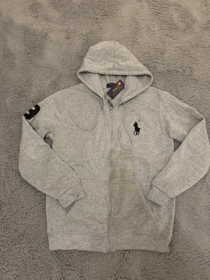 Grå hoodie från Polo Ralph Lauren - Snygg grå hoodie med dragkedja från Polo Ralph Lauren. Klassisk huva med dragsko, svart broderad logga på bröstet och en svart detalj på ärmen. Mjuk bomullsblandning och ribbade muddar för extra komfort.