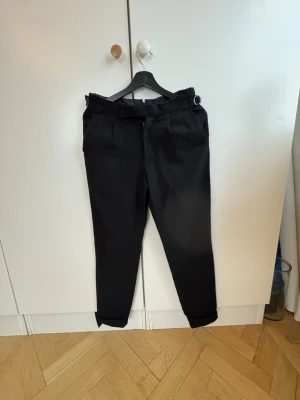  kostymbyxor SUITSUPPLY - Snygga svarta kostymbyxor med hög midja och med spänne på sidorna . Byxorna har uppvikta benslut, sidofickor fram och två bakfickor med knapp. Perfekta för en stilren look och passar till många olika outfits. Köpta på SUITSUPPLY i Stockholm för 2500 kr 