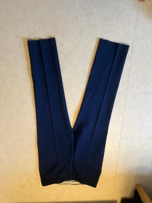 Dressmann kostymbyxa - Snygga mörkblå kostymbyxor med klassiskt pressveck och rak passform. Byxorna har diskret knappstängning i midjan och är tillverkade i ett slätt tyg som ger en stilren look. Perfekta för dig som vill ha en clean och dressad stil.