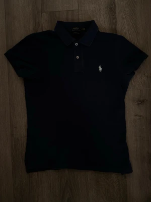Marinblå pikétröja från Ralph Lauren - Snygg marinblå pikétröja från Ralph Lauren med klassisk krage och två knappar framtill. Tröjan har den ikoniska vita logotypen broderad på bröstet och är i en slim passform. Perfekt för en stilren och sportig look. Storlek M men skinny fit så nog mer en S