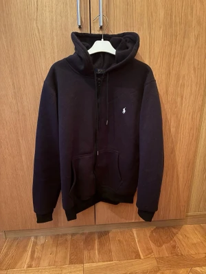 Svart hoodie från Polo Ralph Lauren - Klassisk svart hoodie från Polo Ralph Lauren med dragkedja framtill och vit broderad logga på bröstet. Hoodien har kängurufickor, ribbade muddar och en rejäl huva. Tillverkad i mjukt bomullsmaterial som är skönt att ha på sig.  Säljer åt en kompis som säljer pågrund av att han är för stor för den/ osäker på äkthet därav av ett skit bra pris tveka inte på att skriva om frågor eller om du har funderingar kan gå ner lite i pris vid snabb affär 