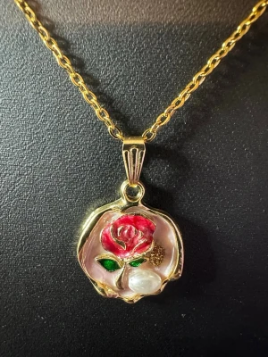 Guldhalsband med ros och pärigt hänge - Unikt halsband med guldfärgad kedja och ett hänge format som en blomma. Hänget har en röd ros med gröna blad, gulddetaljer och en pärlliknande dekoration. Perfekt för dig som gillar romantiska och färgstarka accessoarer.