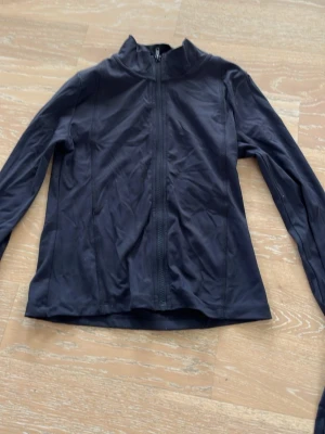 Svart vindjacka med dragkedja - En stilren svart vindjacka med hel dragkedja framtill och hög krage. Jackan har lång ärm och är tillverkad i ett lätt, tunt material som passar perfekt för blåsiga dagar. Enkel och clean design utan synliga loggor eller detaljer.