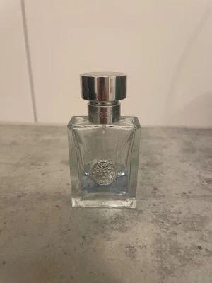 Versace Eau de Toilette Herr - Elegant parfymflaska från Versace med silverfärgad metallkork och transparent glas. Flaskan har en blå tonad vätska och ett klassiskt Versace-emblem på framsidan. Perfekt för dig som gillar exklusiva dofter och stilren design. (OBS 25ml kvar)