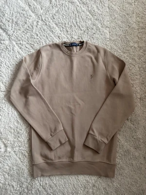 Beige sweatshirt från Ralph Lauren - Säljer en stilren beige sweatshirt från Polo Ralph Lauren med rund halsringning och diskret broderad logga på bröstet. Tröjan har långa ärmar, ribbade muddar och är gjord i mjukt bomullsmaterial. Perfekt för en clean och avslappnad look.