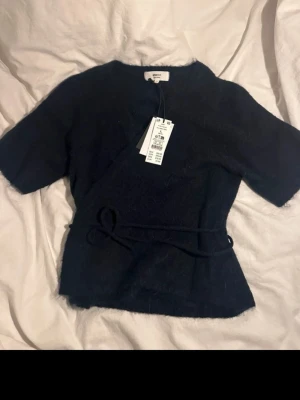 Mörkblå omlottopp från Gina Tricot - Mysig mörkblå omlottopp från Gina Tricot med knytband i midjan och korta ärmar. Toppen har en mjuk, stickad känsla och v-ringad hals. Perfekt för dig som gillar en enkel men trendig look.