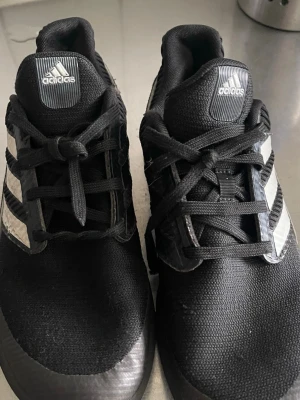 Svarta Adidas sneakers med vita ränder - Snygga svarta sneakers från Adidas med klassiska vita ränder på sidorna. Skorna har svart mesh- och syntetmaterial, samt svart snörning och Adidas-logga på plösen. Perfekta för en sportig och avslappnad stil.