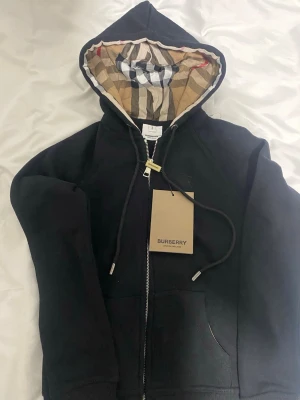 Svart hoodie från Burberry med dragkedja - Säljer en svart hoodie från Burberry med dragkedja och klassiskt rutigt foder i huvan. Hoodien har fickor framtill, justerbar dragsko i huvan och ribbade muddar. Perfekt för dig som gillar stilrena plagg med exklusiva detaljer.
