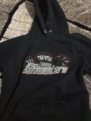 Trapstar London Shooters hoodie svart out - Säljer en svart hoodie från Trapstar med stor broderad 'London Shooters'-logga och puma-tryck på bröstet. Klassisk känguruficka framtill och huva med dragsko. Perfekt för dig som gillar streetwear och vill sticka ut.