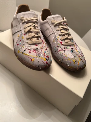 Margiela gats me paint splater  - Unika vita sneakers med coola färgstänk i gult, rött, svart och blått. Skorna har klassisk låg profil, beige snörning och gummisula. Perfekt för dig som vill sticka ut med en kreativ och trendig look. Materialet är textil och lädermix. Säljer då dom va för små för mig 