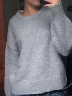 Ljusgrå stickad tröja - Mysig ljusgrå stickad tröja med rund hals och avslappnad passform. Tröjan har ribbstickade muddar vid ärmslut och nederkant. Perfekt för lager på lager och enkel att matcha med jeans eller kjol. Materialet är mjukt och värmande.