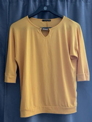 Gul topp med metallspänne - Snygg gul topp från Collection by KappAhl med trekvartsärm och ett unikt metallspänne vid halsringningen. Tillverkad i mjuk bomullsmix och har en avslappnad passform. Perfekt för dig som vill ha något färgglatt och stilrent.
