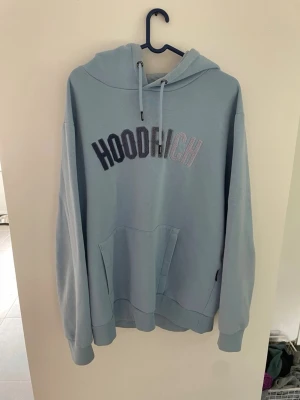 Ljusblå hoodie från Hoodrich - Snygg ljusblå hoodie från Hoodrich med broderad logga i svart och vitt på bröstet. Hoodien har stor huva med dragsko, magficka och ribbade muddar vid ärmar och nederkant. Perfekt för en chill och avslappnad stil.