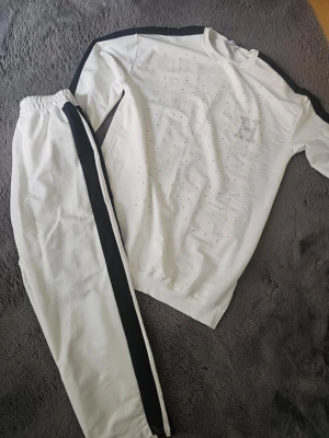 Vitt loungewear-set med svarta detaljer - Snyggt vitt loungewear-set med svarta ränder längs ärmar och byxben. Tröjan har rund hals och är dekorerad med små silverfärgade nitar samt ett stort H på bröstet. Mjukt och bekvämt material, perfekt för chill dagar hemma.