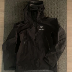  Arcteryx beta ar - Snygg svart vindjacka från Arc'teryx med huva och dragkedja framtill. Jackan har två snedställda fickor med dragkedja och logga på bröstet. Perfekt för blåsiga dagar och outdoor-äventyr. Materialet är tunt och vattenavvisande.