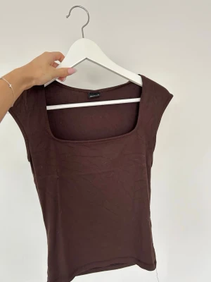 Brun topp med fyrkantig ringning Gina Tricot - Snygg brun topp från Gina Tricot med fyrkantig ringning och ärmlös design. Tillverkad i mjuk bomull som sitter skönt mot kroppen. Perfekt att styla med jeans eller kjol för en trendig look.