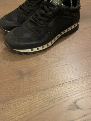 Svarta Valentino sneakers med nitar - Snygga svarta sneakers från Valentino med coola nitar längs sulan och diskret camomönster i textil och mocka. Skorna har svarta skosnören och en vit mellansula med silverfärgade nitar som ger en unik look.