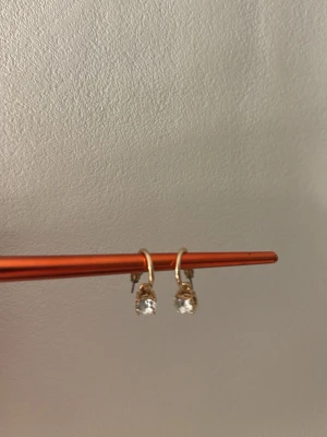 Guldtonade örhängen med kristallhänge, nickelfria - Snygga örhängen i guldtonad metall med runda, klara kristallhängen som ger en elegant touch. Perfekta för att lyfta vilken outfit som helst med lite extra bling. Klassisk design med enkel ring och gnistrande sten.