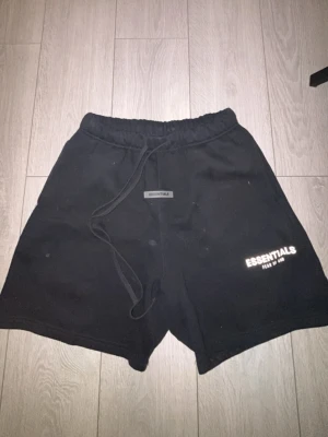 Svarta Essentials shorts med snörning - Svarta shorts från Essentials med elastisk midja och snörning. Logotypen är tryckt på framsidan och det finns en bakficka. Perfekta för chill eller träning, tillverkade i mjukt material som känns skönt mot huden.