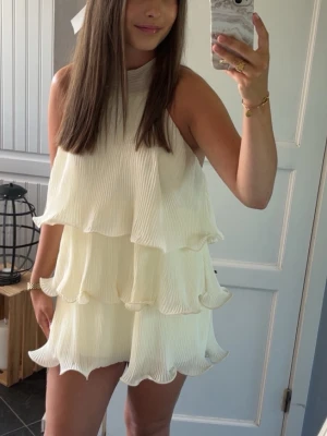 Söt klänning - Säljer en ljusgul kortklänning med flera lager volanger och hög hals. Klänningen är ärmlös och har ett luftigt, plisserat tyg som ger en snygg och modern look. Perfekt för sommarens alla tillfällen. 