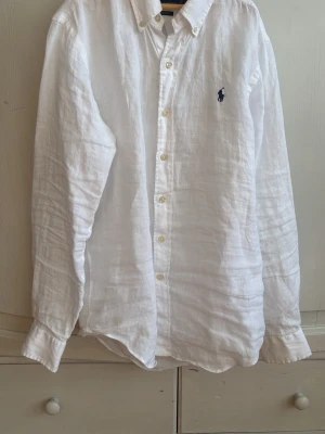 Vit linneskjorta från Polo Ralph Lauren - Klassisk vit skjorta i linne från Polo Ralph Lauren med button-down krage och broderad logga på bröstet. Skjortan har långa ärmar och knäppning framtill. Perfekt för en clean och stilren look.