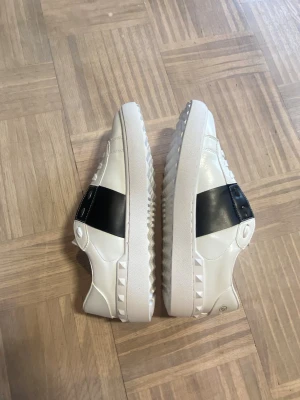 Vita sneakers med svart detalj - Snygga vita sneakers från Valentino med bred svart läderdetalj över sidan och nitar längs sulan. Skorna har klassisk låg profil, vita skosnören och en chunky sula. Perfekta för dig som gillar stilrena men ändå unika sneakers.