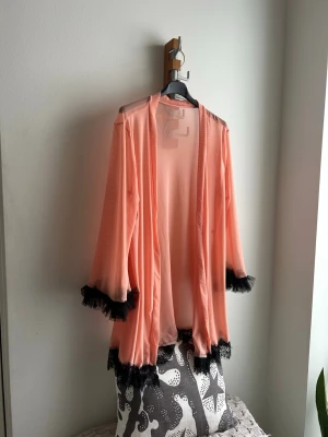 Mmorgonrock med spets - Säljer en super fin morgonrock med svarta spetsdetaljer och volanger vid ärmslut och nederkant. Morgonrocken har vida ärmar och är tillverkad i ett tunt, mjukt tyg som känns lyxigt och skönt. Helt ny och aldrig använd!