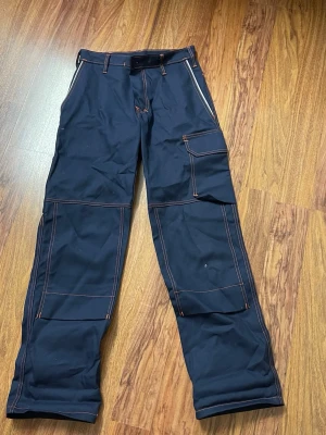 Arbetsbyxor/Blåa cargobyxor med orange sömmar - Snygga blåa cargobyxor med kontrasterande orange sömmar och flera praktiska fickor på benen. Byxorna har bälteshällor och raka ben, perfekta för en streetwear-look. Materialet känns slitstarkt och passar till dig som gillar funktionella detaljer.