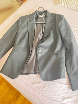 Klassisk svart kavaj med en knapp - Stilren svart kavaj med en knapp framtill och klassisk krage. Kavajen har två fickor utan lock och är helfodrad med slät insida. Passformen är rak och materialet ser ut att vara en blandning av polyester och viskos.