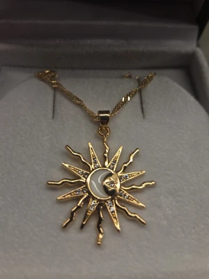 Guldigt halsband med sol och ädelstenar - Unikt halsband i guldton med ett hängsmycke som föreställer en sol och måne. Solstrålarna är dekorerade med små gnistrande stenar och månen har en ansiktsdetalj. Kedjan är tvinnad och ger an young och trendig vibe. Perfekt för dig som gillar statement-smycken och set med astrologiska motiv.