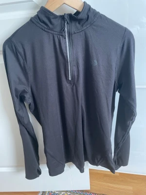 Svart långärmad träningströja lager 157 - Svart långärmad träningströja med halv dragkedja framtill och hög krage. Tillverkad i ett stretchigt funktionsmaterial som passar perfekt för löpning eller gym. Tröjan har en enkel och stilren design utan synliga loggor.