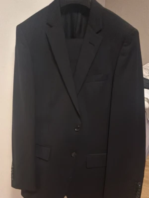 Hugoboss kostym slim fit set.  - Hugo Boss kostym för herr i svart ull, storlek 46 på kavajen och storlek 48 på byxan. Kostymen består av kavaj och byxa i matchande tyg. Kavajen har klassisk krage med slag, tvåknäppning framtill samt fickor med lock. Byxorna är raka i modellen med pressveck. Kavajen i hög kvalitet  med yttertyg i 100% ull, foder i 100% cupro och ärmfoder i 50% cupro/50% viskos, vilket ger en mjuk och bekväm känsla samt bra andningsförmåga. Nypris 8000kr. Bilderna gör tyvärr inte kostymen helt rättvisa. 