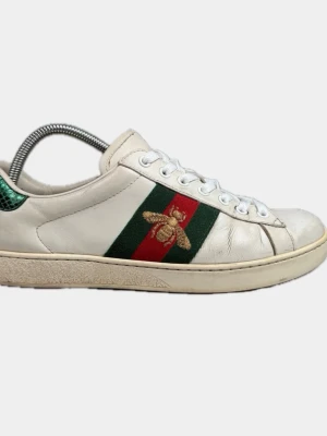 Gucci Ace Bee vita sneakers - Gucci Ace sneakers i vitt läder med klassiska gröna och röda ränder på sidan samt broderad guldfärgad bi. Grön detalj i ormskinnsmönster på hälen. Låga sneakers med vita skosnören och gummisula. 
