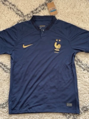 Frankrike fotbollströja Nike blå - Mörkblå fotbollströja från Nike med Frankrikes landslagslogga och två stjärnor på bröstet. Tröjan har korta ärmar, guldfärgade detaljer och klassisk krage med knapp. Perfekt för dig som älskar fotboll och vill visa ditt stöd för Les Bleus.