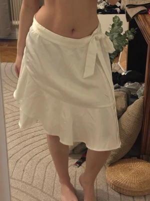 Vit omlottkjol med volang - Snygg vit omlottkjol med volangkant och knyt i midjan. Kjolen har en lätt glansig yta och är perfekt för dig som gillar en feminin och fräsch stil. Passar till både sneakers och sandaler.