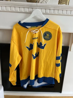 Gul Tre Kronor hockeytröja Reebok - Säljer en gul hockeytröja från Reebok med Tre Kronor-logga och blå detaljer. Tröjan har snörning i halsen, blå muddar och ränder på ärmarna samt ett märke på bröstet. Perfekt för dig som vill stötta Sverige på match eller träning. Barn Size 160/170