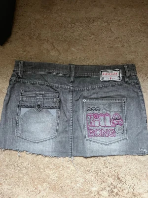 Grå jeanskjol från Billabong - Säljer en grå kort jeanskjol från Billabong med rå kant nertill, flera fickor och en cool dragkedjedetalj fram. Baksidan har broderad Billabong-logga i rosa och vitt. Perfekt för dig som gillar streetwear och denim.