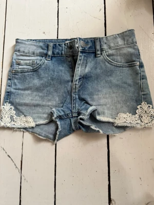 Blå jeansshorts med spetsdetaljer - Snygga blå jeansshorts med rå kant och vita spetsdetaljer på sidorna. Klassisk femficksmodell med knapp och dragkedja framtill. Perfekta för sommaren och ger en avslappnad men ändå söt look.