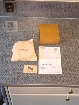 Burberry bälte med silverfärgat spänne - Snyggt Burberry bälte med klassiskt rutigt mönster på insidan och svart utsida. Bältet har ett stort, silverfärgat burberry spänne och levereras med originalpåse, box och certifikat. Perfekt accessoar för att lyfta din outfit.