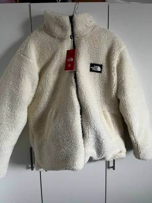 Reversible pufferjacka från The North Face - Säljer en fluffig och varm vit och blå pufferjacka från The North Face. Jackan har mjukt teddy-material på utsidan, dragkedja framtill och två sidofickor. Loggan sitter broderad på bröstet. På andra sidan är den ljusblå. Perfekt för kalla vinterdagar och väldigt bra att ha två jackor i en. Säljs då storleken inte passar M mer L. Helt ny oanvänd med tagg på.🩵