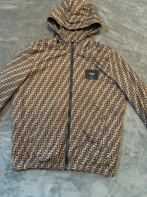 Fendi vindjacka med FF-mönster - Snygg vindjacka från Fendi med ikoniskt brunt och svart FF-logomönster över hela jackan. Jackan har huva, hel dragkedja framtill och en svart patch med Fendi-logga på bröstet. Tillverkad i lätt och smidigt material, perfekt för streetwear och trendiga looks. Det är Storlek L men det passar M också.