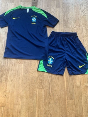 Brasilien fotbollsträningsset Nike - Säljer ett blått och grönt Brasilien fotbollsträningsset från Nike. Setet består av en kortärmad tröja och shorts, båda med Brasilien-logga och Nike swoosh i gult. Perfekt för träning eller match. Materialet är lätt och andas, designat för sport.